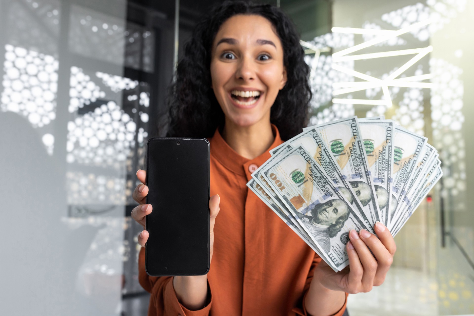 Businesswoman happy happy with winning shows blank smartphone screen to camera and holds money American cash dollars in hands, Hispanic woman inside office at workplace